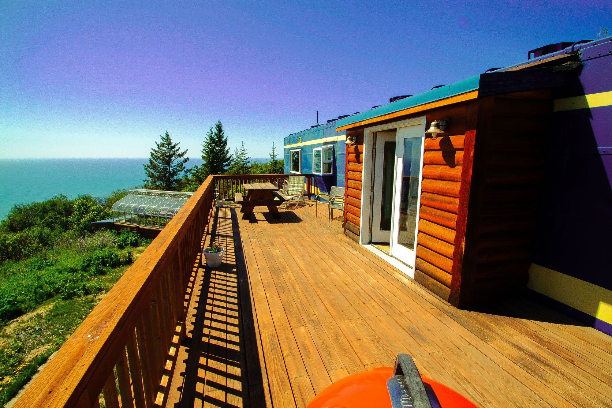 A wooden deck at one of the cabins in Homer, Alaska, overlooking the ocean with a picnic table, sliding glass doors, and a partial view of a greenhouse below.