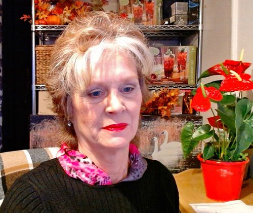 A woman with light hair wearing a black sweater and a patterned scarf sits beside a potted red anthurium plant at Alaska Adventure Cabins.