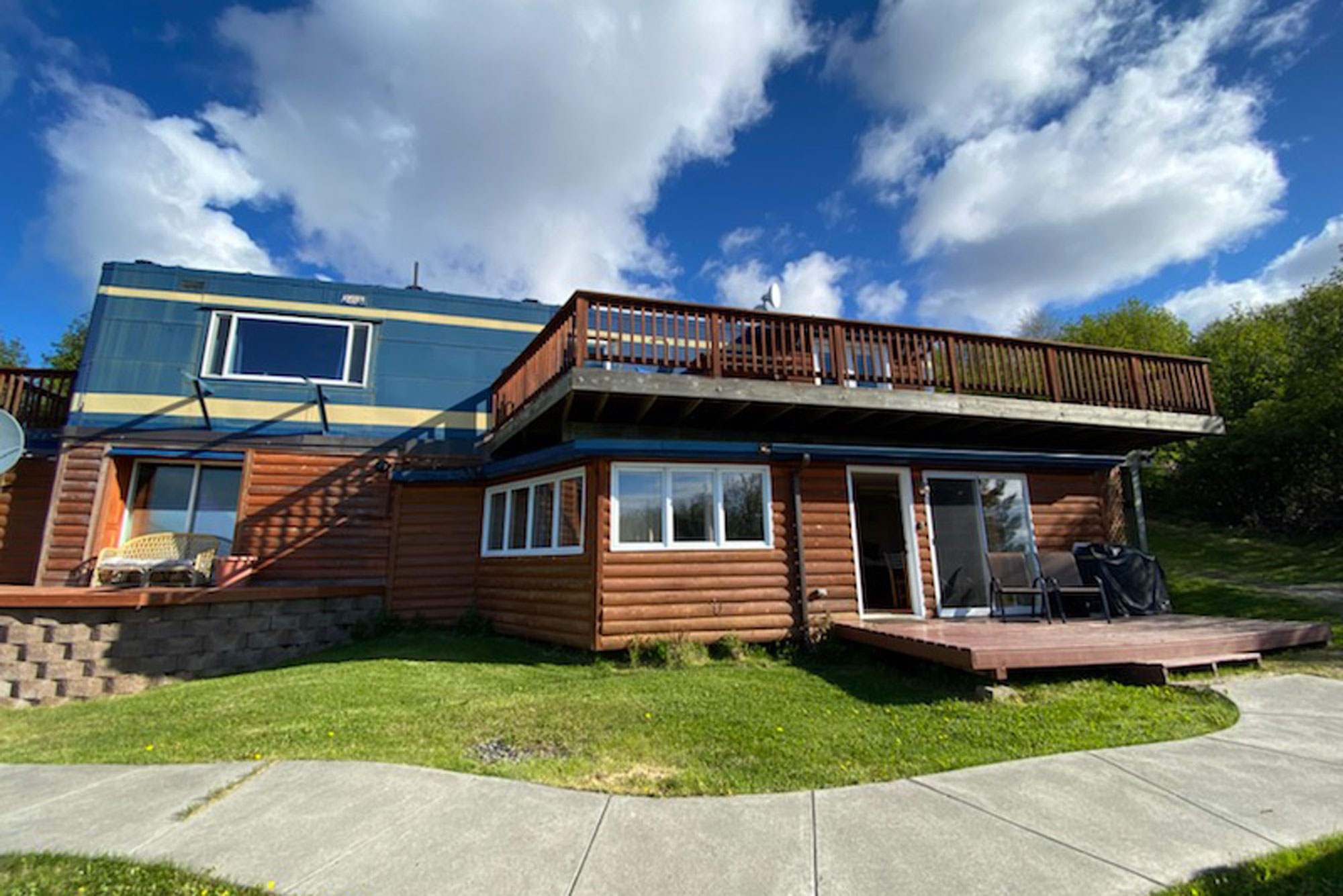 A two-story log cabin in Homer, Alaska, featuring a large wooden deck on the upper level and a concrete walkway in the foreground.
