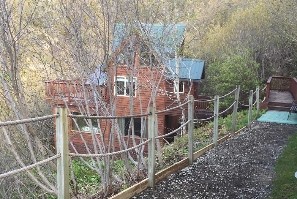 A wooden cabin with a green roof nestled in a wooded ravine in Homer, Alaska, viewed from a gravel path with a rope railing.
