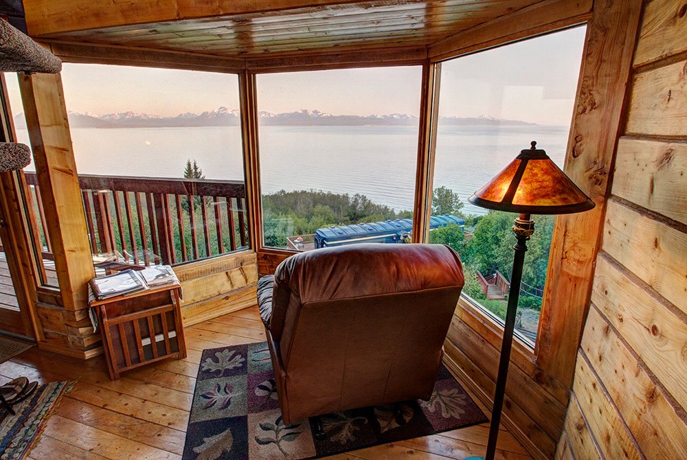 A brown leather armchair in a cabin in Homer, Alaska, faces a large bay window overlooking a calm body of water with distant snow-capped mountains.