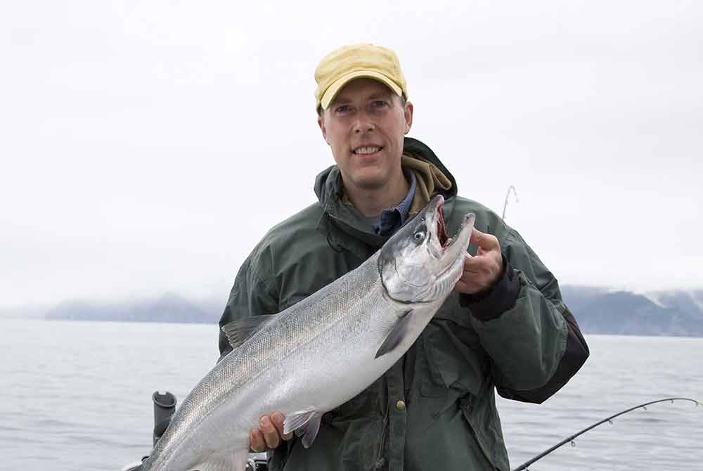 A man in a yellow cap and green jacket holds a large salmon on a boat near cabins in Homer, Alaska.